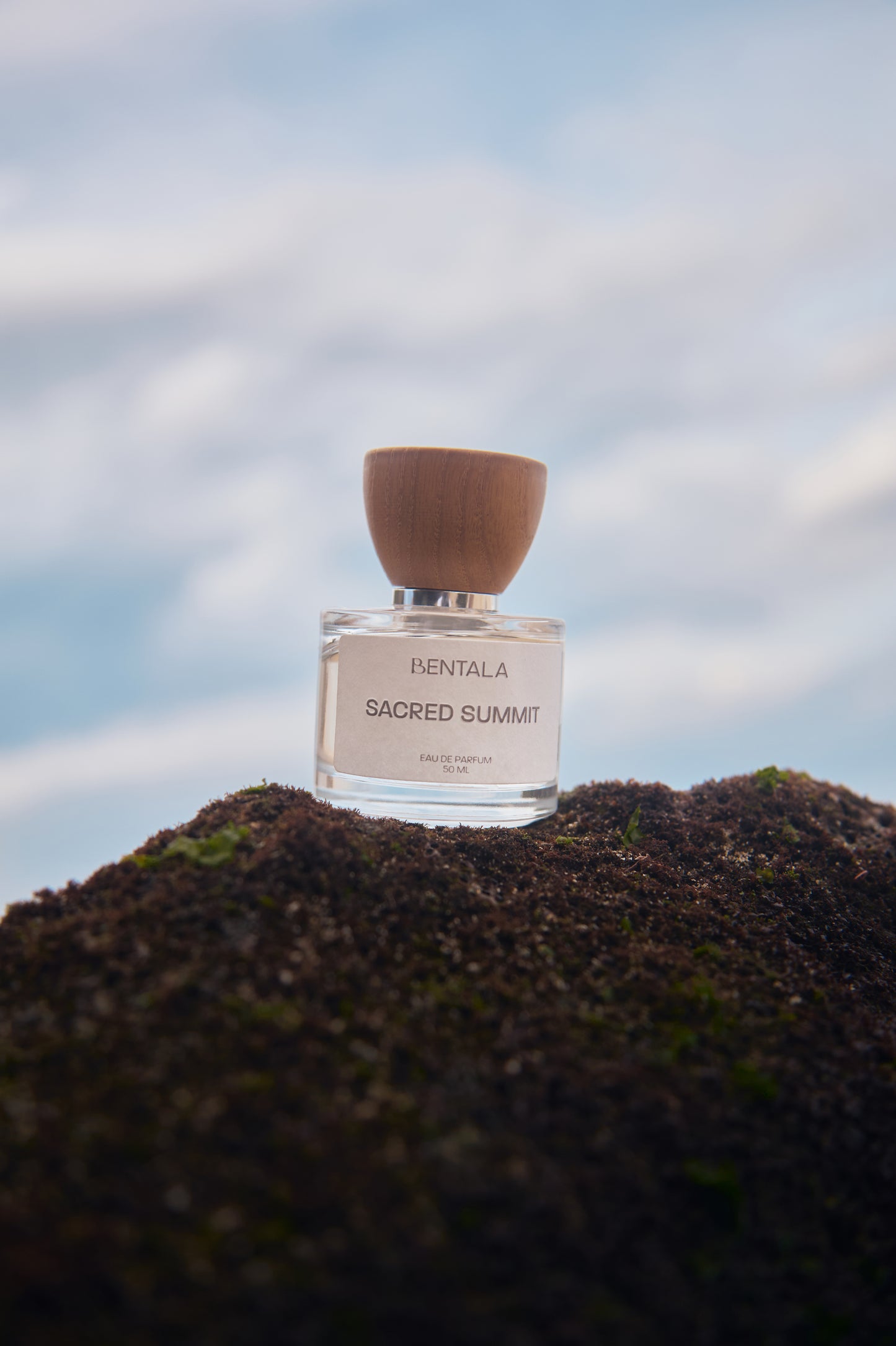 Perfume bottle labeled 'Bentala Sacred Summit' on a Rocky Mountain with a blurred sky background