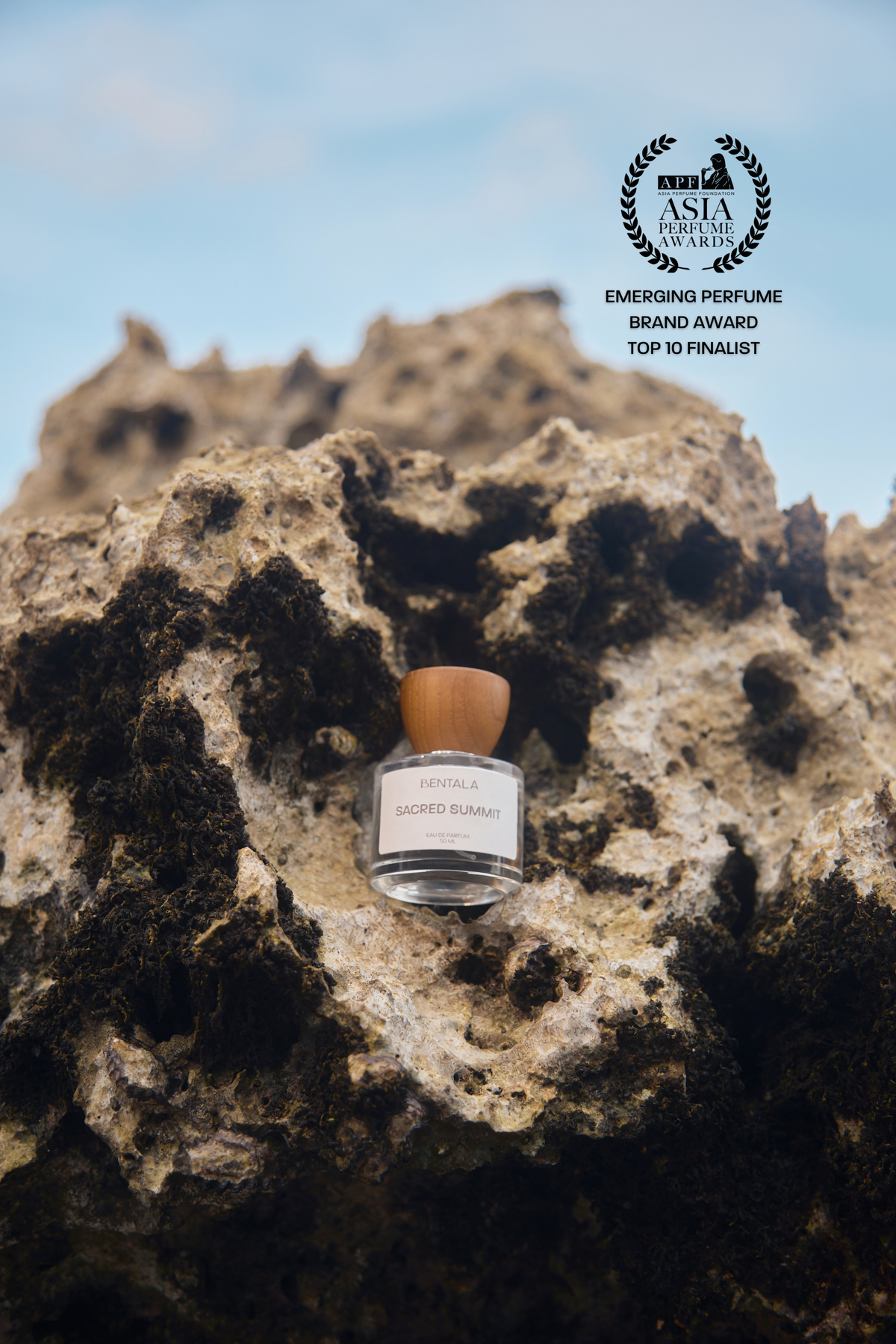 A small perfume bottle labeled 'Scent of Arabia' with a light brown cap, placed on a dark textured rock with a blue sky background. An award badge reading 'Emerging Perfume Brand Award Top 10 Finalist' appears in the upper right.